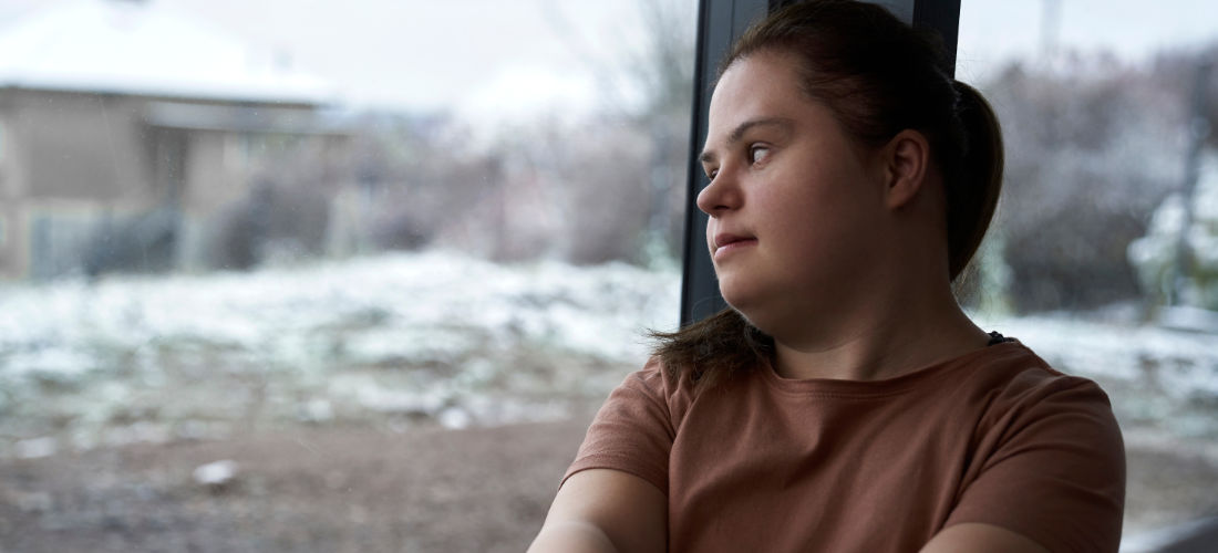 Eine junge weiße Frau mit Down Syndrom schaut nachdenklich aus dem Fenster.