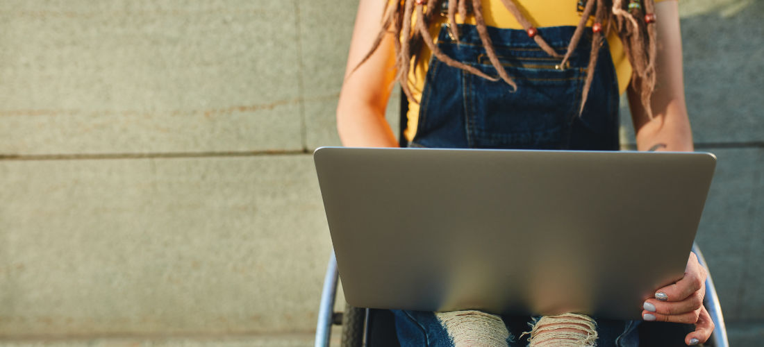 Eine weiße junge Rollstuhlfahrerin hat einen großen Laptop auf dem Schoß aufgeklappt.