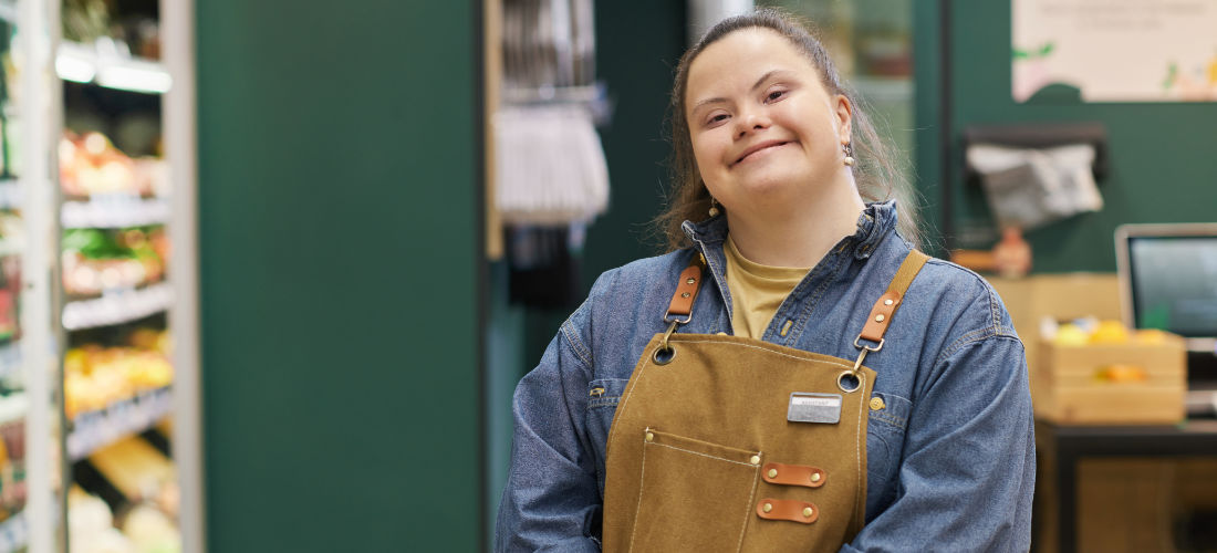 Eine junge weiße Mitarbeiterin mit Downsyndrom im Supermarkt im Kassenbereich lächelt.