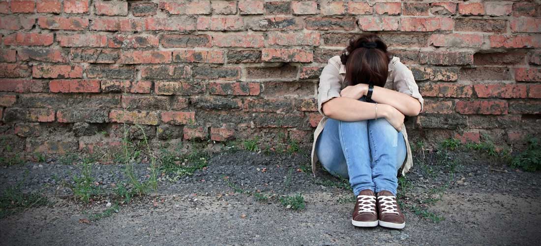 Mädchen sitzt vor einer Mauer. Die Mauer ist aus Ziegel-Steinen. Das Mädchen hat ihren Kopf auf die Knie gelegt.