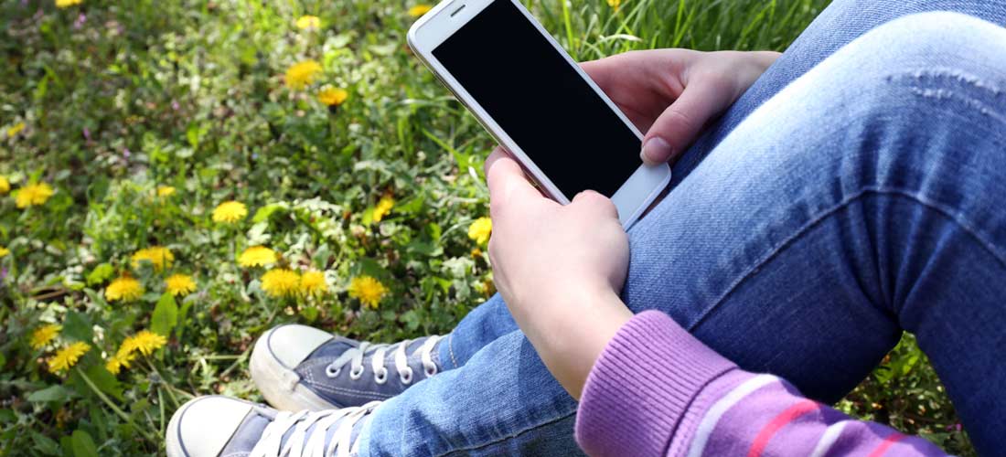 Bildausschnitt von sitzender Jugendlicher auf Löwenzahn-Wiese, mit ihren Armen um ihre Knie und Smartphone in der Hand.