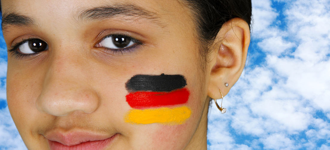 Portrait-Ausschnitt von Mädchen mit dunklen Augen, Deutschlandfahne auf Wange geschminkt, vor blauem Himmel.