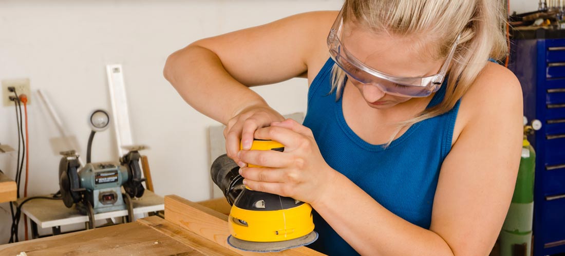 Mädchen mit Schutz-Brille. Sie hat ein Gerät in der Hand. Und schleift mit dem Gerät Holz ab.