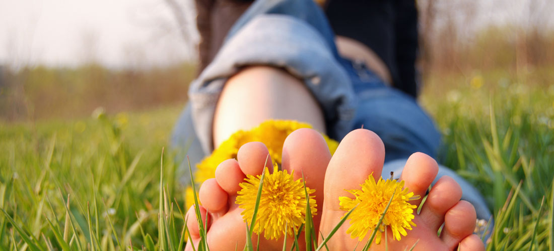 Jugendliche sitzt auf Wiese, Barfußsohlen sichtbar zwischen deren größten Zehen jeweils eine Löwenzahn-Blüte steckt.