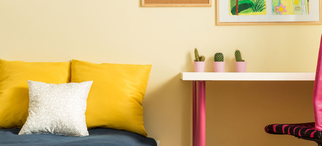 Ein Ausschnitt von einem Zimmer. Die Farbe der Wand ist gelb. Es gibt einen Schreibtisch. Der Schreibtisch ist weiß-rosa. Es gibt einen Schreibtisch-Stuhl. Der Schreibtisch-Stuhl ist rosa. Es gibt Sofa-Kissen. Es gibt ein Bild. Und es gibt eine Pinnwand.