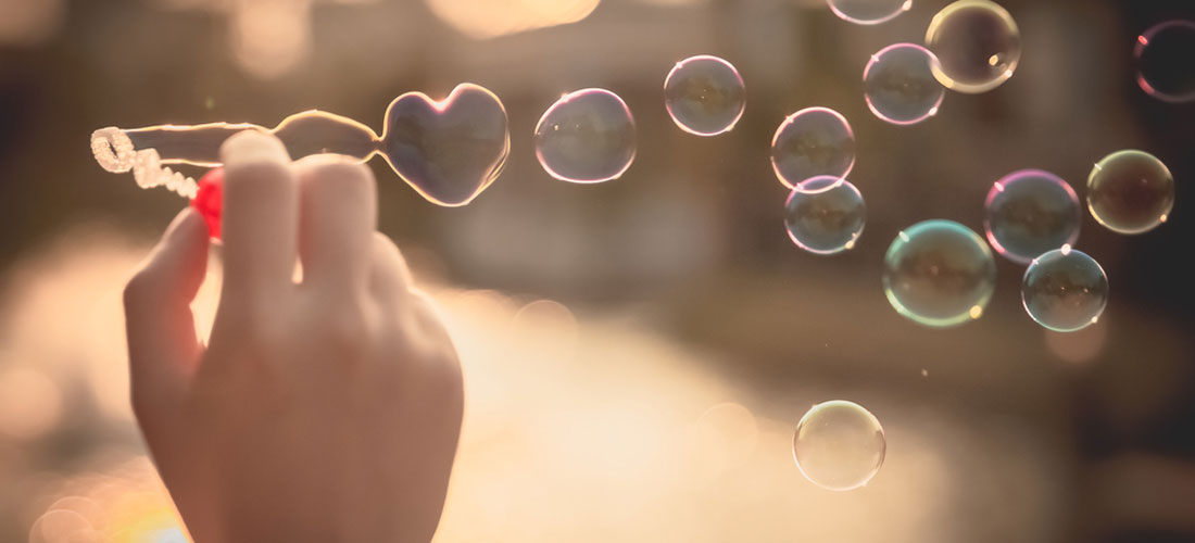 Eine Hand hält Seifen-Blasen-Stäbchen. Aus dem Seifen-Blasen-Stäbchen kommen Seifen-Blasen. Eine Seifen-Blase in Herz-Form. Alle Seifen-Blasen fliegen in der Luft.