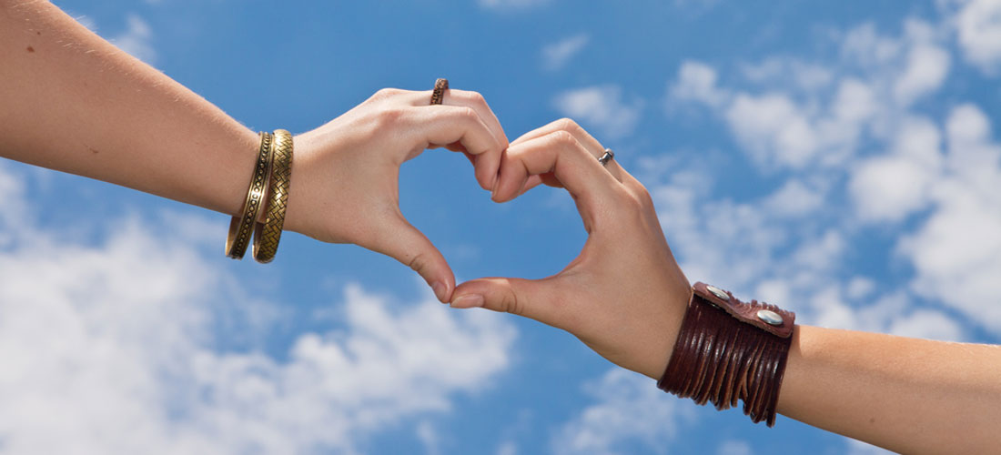 Zwei unterschiedliche Hände bilden ein Herz. Das Herz steht in der Luft. Im Hintergrund blauer Himmel.